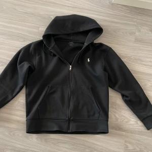 Svart hoodie från Polo Ralph Lauren - Säljer en stilren svart hoodie från Polo Ralph Lauren med dragkedja och huva. Den har en liten logotyp på bröstet och praktiska fickor framtill. Perfekt för en avslappnad look.
