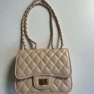 Beige quiltad axelväska - Snygg beige axelväska med quiltat mönster och kedjeaxelrem. Väskan har en praktisk låsdetalj framtill och är perfekt för våren! Kontakta för frågor💕