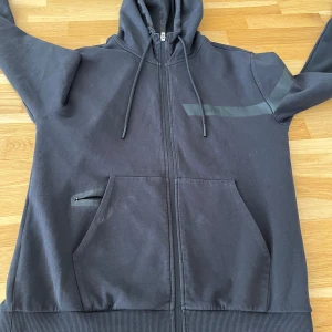 Svart hoodie från Boss - Säljer en stilren svart hoodie från Boss med dragkedja och justerbar huva. Den har en diskret logga på bröstet och gröna detaljer vid kragen. Perfekt för en avslappnad look. Kan även byta !