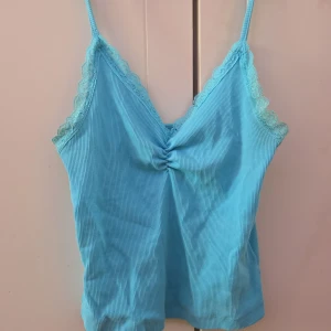 Turkos ribbad linnetopp med spetsdetaljer - Säljer en söt turkos ribbad linnetopp med smala axelband och spetsdetaljer längs kanterna. Toppen har en knytdetalj framtill som ger en fin passform. Perfekt för varma sommardagar eller som ett lager under en kofta.