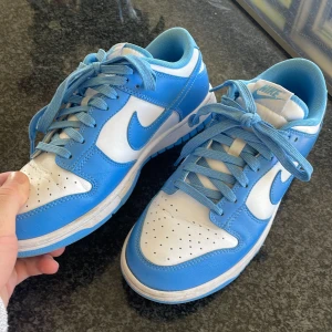 Nike Dunk UNC - Snygga Nike Dunk sneakers i en fräsch blå och vit färgkombination. Säljs då jag har växt ur dom. Sitter riktigt bekvämt och är en perfekt sko till sommaren! Lådan är borttappad tyvär.