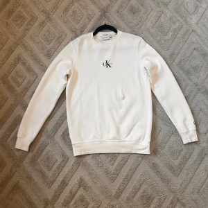 Vit sweatshirt från Calvin Klein - Säljer en stilren vit sweatshirt från Calvin Klein med deras ikoniska CK-logga på bröstet. Tröjan har långa ärmar och en klassisk rund halsringning. Perfekt för en avslappnad och trendig look.