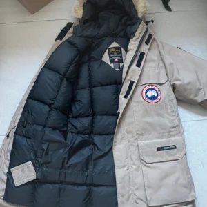 Beige dunjacka från Canada Goose - Säljer en beige dunjacka från Canada Goose med svart foder och en ikonisk logotyp på ärmen. Jackan har en pälsfodrad huva och flera praktiska fickor. Perfekt för kalla vinterdagar. MVH EliteVolt