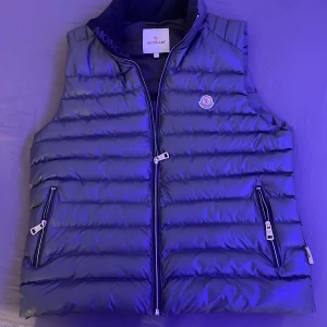 Grå dunväst från Moncler - Säljer en snygg blå dunväst från Moncler med dragkedja och två praktiska fickor framtill. Västen har Monclers logotyp på bröstet och är perfekt för kyligare dagar. Den är lätt och bekväm att bära.