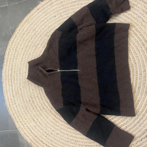 Brun och svart randig tröja med dragkedja - Säljer en mysig brun och svart randig tröja med dragkedja framtill. Tröjan har en hög krage och långa ärmar, perfekt för kyligare dagar. Den är både stilren och bekväm.