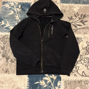 Svart hoodie från Sail Racing - Snygg svart hoodie från Sail Racing med dragkedja och huva. Tillverkad i en bomullsblandning för extra komfort. Perfekt för en avslappnad stil. St 160 
