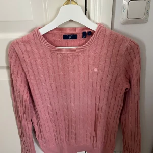 Rosa stickad tröja från Gant - Säljer en snygg rosa stickad tröja från Gant med kabelstickat mönster. Tröjan har långa ärmar och en rund halsringning. Perfekt för kyligare dagar. 🧶