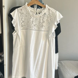 Vit blus med spetsdetaljer från Mohito - Säljer en elegant vit blus från Mohito, köpt i Polen.  vackra spetsdetaljer på framsidan. Blusen har korta ärmar och en rund halsringning med volangkant. Perfekt för en stilren look. Använd 1 gång. Nypris: 300kr