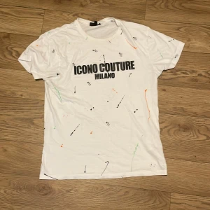 Vit t-shirt från Icono Couture - Säljer en vit t-shirt från Icono Couture med texten 'ICONO COUTURE MILANO' i svart på framsidan. T-shirten har ett unikt stänkmönster i olika färger som ger en cool touch. Perfekt för dig som vill sticka ut!