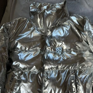 Silver pufferjacka från Alexander Wang - Säljer en cool och glansig silverfärgad pufferjacka från Alexander Wang. Jackan har en hög krage och dragkedja framtill. Perfekt för att sticka ut i mängden med sin futuristiska look. 🛍️✨