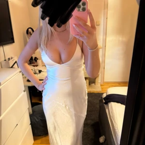 Vit satin klänning - Snygg vit satinklänning med smala axelband och öppen rygg. Klänningen har en elegant slits och knytning i ryggen för en justerbar passform. Perfekt som balklänning eller till skolavslutning!