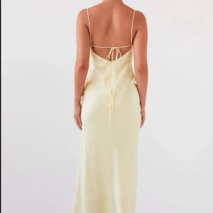 Ljusgul långklänning i satin - Elegant ljusgul långklänning, från Peppermayo, i satin med smala axelband och öppen rygg. Klänningen har en drapering framtill och knytning bak för en justerbar passform. Perfekt för sommarens festligheter. Köpt för 814kr