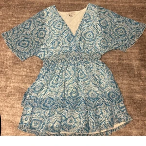 Blå och vit mönstrad klänning - Säljer en blå och vit kortklänning med ett unikt mönster. Klänningen har en v-ringad design och korta ärmar. Perfekt för sommarens varma dagar!