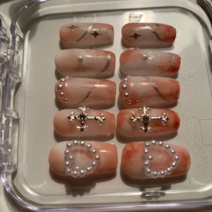 Naglar med pärlor och korsdetaljer - Snygga lösnaglar i en mjuk orange nyans med silverglittriga detaljer. Dekorerade med pärlor, kors och hjärtan för en unik look. Perfekt för att ge dina händer en extra touch av glamour.