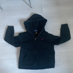Svart hoodie från Ralph Lauren - Säljer en klassisk svart hoodie från Ralph Lauren med dragkedja och huva. Den har en liten röd broderad logga på bröstet. Perfekt för en stilren och bekväm look.