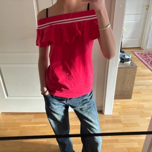 Röd offshoulder topp med volang  - Säljer en snygg röd offshoulder topp med volangdetaljer och vita ränder. Perfekt för en somrig look. Toppen är ärmlös och har en avslappnad passform.