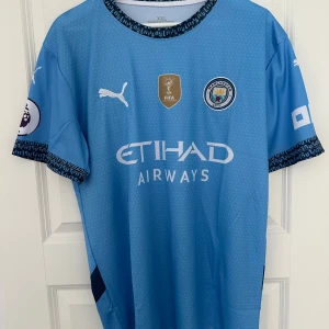 Blå Manchester City fotbollströja från Puma - Säljer en blå Manchester City fotbollströja från Puma med korta ärmar. Tröjan har klubbens emblem och sponsorlogga på framsidan samt spelarnamn och nummer 47 på baksidan. Perfekt för fans av laget!
