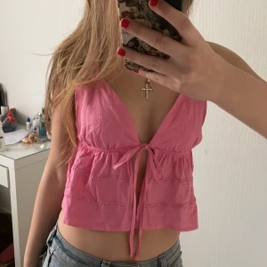 Rosa ärmlös topp med knyt - Säljer en söt rosa ärmlös topp med knyt framtill. Toppen har en v-ringad design och är perfekt för varma dagar. Den är lätt och luftig, vilket gör den idealisk för sommaren.