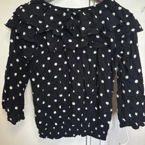 Svart prickig blus med volang - Säljer en svart blus med vita prickar och bred volang runt axlarna. Blusen har offshoulder-design och långa ärmar. Perfekt för en trendig look.🩷