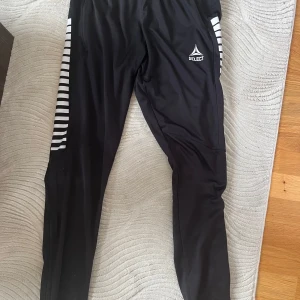 Svarta träningsbyxor från Select L - Svarta träningsbyxor från Select i storlek L, tillverkade i 100% polyester. Byxorna har en slim passform och är perfekta för träning. Elastisk midja för skön komfort och har dragkedjor för fickor och öppning vid benets slut. Inga skador, endast små slitningar på trycket.