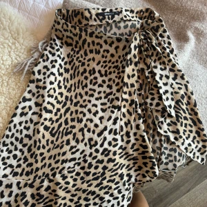 Leopardmönstrad omlottkjol Gina Tricot - Säljer en snygg omlottkjol från Gina Tricot i storlek 34 med leopardmönster. Kjolen har beige och svarta toner och knyts i sidan för en cool look. Perfekt för dig som vill sticka ut med ett djuriskt mönster och trendig stil.