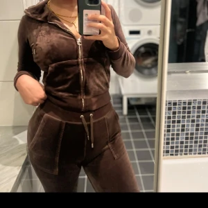 Juicy Set - Säljer en brun juicy set med dragkedja framtill och huva. Tröjan har långa ärmar och fickor på sidorna, perfekt för chill dagar. Snyggt guldigt detaljer på dragkedjan och snörning vid midjan. Passar dig som gillar comfy och trendigt.