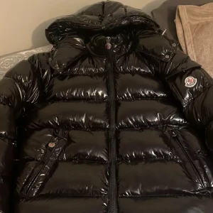 Svart glansig dunjacka från Moncler - Säljer en svart, glansig dunjacka från Moncler med klassisk pufferstil och huva. Jackan har dragkedja framtill, två fickor med tryckknappar och ikoniskt Moncler-märke på ärmen. Perfekt för kalla dagar när du vill hålla stilen och värmen.