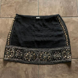 Svart minikjol med paljetter och pärlor  - Supersnygg svart minikjol från Bik Bok med detaljerade guldpaljetter och pärlor längs kanterna och nederdelen. Kjolen har ett lyxigt broderat mönster med metallic och pärlbroderier som ger en unik vibe. Perfekt för dig som gillar statement-plagg och vill sticka ut. Aldrig använd, storlek M, normal i storleken.