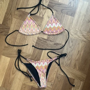 Färgglad bikini med snörning - Säljer en snygg bikini med färgstarkt mönster i rosa, orange, gul och vit. Bikinin har trekantsformad överdel och smala svarta snören som knyts i nacke, rygg och på sidorna av underdelen. Materialet är mjukt och stretchigt, perfekt för strandhäng och pool.