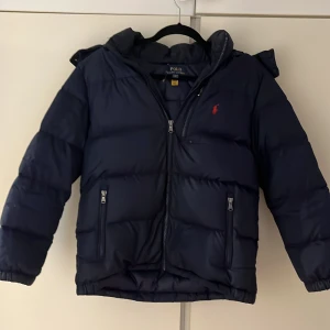 Mörkblå pufferjacka Polo Ralph Lauren - Snygg mörkblå pufferjacka från Polo Ralph Lauren med huva och två dragkedjeförsedda fickor. Jackan har det klassiska röda Polo-logot på bröstet och är tillverkad i polyester. Perfekt för kalla dagar och har en modern, loose passform. Men jackan har lite små hål som inte syns när du har på dig den.