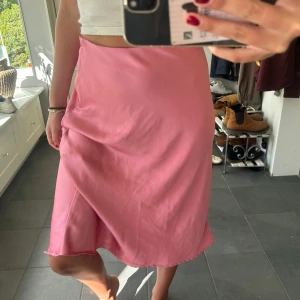 Rosa omlottkjol i satin - Säljer en superfin rosa omlottkjol med lätt glansig satinfinish. Kjolen har en mjuk och flowy passform som ger en snygg siluett. Perfekt för dig som gillar färg och vill ha en kjol som känns både trendig och bekväm.