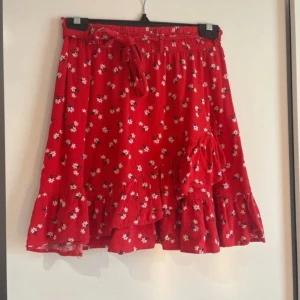 Röd kjol med vita blommor - Säljer en röd kjol med vita blommor och volang nertill från Lindex. Kjolen är i storlek 158/164 (barnstorlek) men funkar också för storlek XXS och XS.  Mycket använd men i väldigt bra skick.