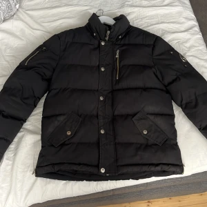 Svart pufferjacka Moose Knuckles - Svart pufferjacka från Moose Knuckles med broderad logga på insidan och metallpatch på ärmen. Jackan har hög krage, flera fickor med dragkedja och knappar, samt quiltad design. Perfekt för kalla dagar och har en cool streetstil.