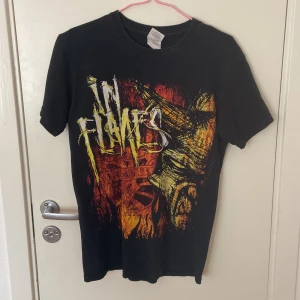 In Flames band t-shirt Gildan S - Svart band t-shirt från Gildan med fet In Flames-tryck fram och turnédatum på ryggen. Grym design i rött, gult och orange med coolt motiv och text. Gjord i 100% bomull, klassisk passform och korta ärmar. Perfekt för dig som älskar metal och vill sticka ut.