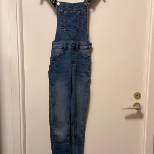Blå jeansbyxdress från H&M, strl 32 - Säljer en snygg blå jeansbyxdress från H&M i storlek 32. Byxdressen har klassiska justerbara axelband med metallspännen, en stor ficka på bröstet och flera fickor framtill. Materialet är jeans med en cool tvättad look som ger en avslappnad vibe.