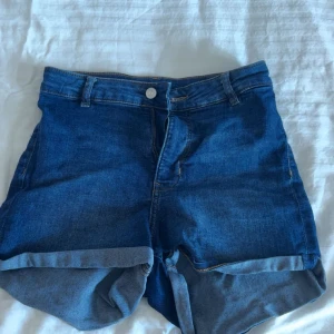 Blå högmidjade jeansshorts - Snygga blå jeansshorts med hög midja och uppvikta benslut. Klassisk femficksmodell med knapp och dragkedja framtill. Perfekta för varma dagar och passar till allt. Materialet är slitstarkt denim som ger en cool och avslappnad vibe.