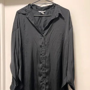 Svart oversized skjorta i silke - Snygg svart skjorta med oversized passform och klassisk krage. Tillverkad i mjukt och glansigt silke, perfekt för dig som gillar en chill och stilren look. Skjortan har långa ärmar och knappar framtill. Enkel att matcha med jeans eller kostymbyxor.