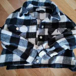 Rutig schakets från Zara - Säljer en snygg schakets från Zara i svart och vit rutig design. Jackan har krage, tryckknappar framtill och två bröstfickor med lock. Materialet är mjukt och känns som ull, perfekt för lager på lager. Passar dig som gillar streetstyle och chill vibe.