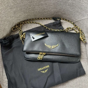 Svart axelväska från Zadig & Voltaire - Snygg svart axelväska i skinn från Zadig & Voltaire med guldfärgade detaljer och kedja. Väskan har ett coolt guldigt vingemblem på framsidan och dubbla dragkedjor. Perfekt storlek för det viktigaste och ger en lyxig vibe till din outfit.