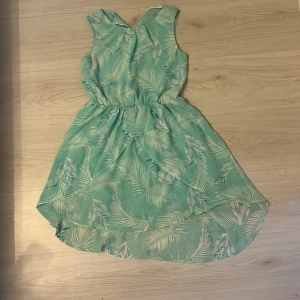 Mintgrön ärmlös klänning från H&M - Superfin mintgrön klänning från H&M med tropiskt bladmönster. Modellen är ärmlös och har en lätt rundad halsringning samt resår i midjan för en snygg siluett. Materialet är tunt och luftigt, perfekt för varma dagar. Baksidan har en liten v-ringad detalj.