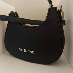 Svart axelväska från Valentino - Snygg svart axelväska från Valentino med guldfärgade detaljer och dragkedja upptill. Väskan har en stilren, rundad form och är tillverkad i skinn med ett diskret texturerat ytskikt. Perfekt storlek för det viktigaste och har ett elegant metallogo framtill. Säljs på grund av ingen användning 