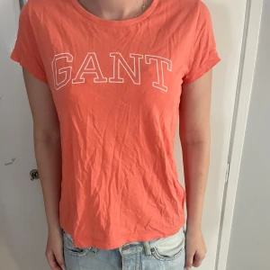 Korallfärgad GANT t-shirt i bomull - Snygg korallfärgad t-shirt från GANT med vit logotyp framtill. T-shirten har rund hals, korta ärmar och är gjord i mjuk bomull. Passformen är normal och den är perfekt för en chill och avslappnad stil.