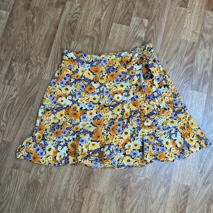Blommig kjol från HM - Blommig kort kjol från HM Divided i storlek S. Kjolen har ett färgglatt mönster med gula, lila och vita blommor, elastisk midja och en söt knyt-detalj framtill på ena sidan. Rosetten går att justera. Använd en gång.