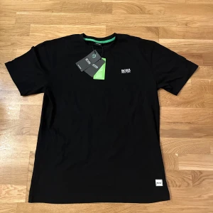 Svart t-shirt från Hugo Boss - Svart t-shirt från Hugo Boss med vit logotyp på bröstet och en liten etikett vid nederkanten. Klassisk rund hals med grön insida och korta ärmar. Tillverkad i mjuk bomull som känns skön mot huden. Perfekt för dig som gillar stilrena och enkla plagg.