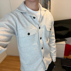 Ljusgrå overshirt från Zara - Säljer en stilren ljusgrå overshirt från Zara med klassisk krage och svarta knappar. Två stora bröstfickor med lock och knapp framtill. Materialet är mjukt och känns som ullblandning, perfekt för lager-på-lager. Passar dig som gillar clean och enkel stil.