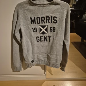 Grå stickad tröja Morris Gents XS - Snygg grå stickad tröja från Morris Gents i storlek XS. Tröjan har svart broderad text och emblem på bröstet, samt ribbade muddar vid ärmslut och nederkant. Perfekt för dig som gillar klassisk stil med en modern touch. Priset ej hugget I sten.