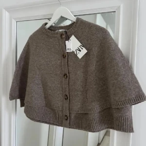 Beige poncho Zara  - Superfin beige stickad poncho från Zara med knappar hela vägen framtill. Modellen är loose och har korta ärmar, perfekt att slänga över en t-shirt eller långärmad. Ribbad kant nedtill och runt halsen ger en chill och snygg look. Ponchon har inte lappen kvar som på första bilden, men är ändå i nyskick.