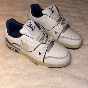 Louis Vuitton LV Trainer sneakers vit/blå - Säljer ett par Louis Vuitton LV Trainer sneakers i vitt och marinblått med beige detaljer. Skorna har snörning, rund tå och platt sula. Ikoniska LV-loggor syns på tungan och sidan, samt #54 på hälen. Tillverkade i skinn med snygga kontrastdetaljer och mönstrad yttersula.