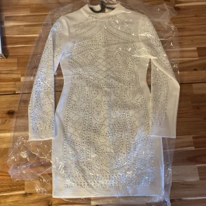 Vit kort klänning med silverdetaljer - Säljer en vit kort klänning med lång ärm och glittriga silverpaljetter i ett snyggt mönster framtill och på ärmarna. Klänningen har en figurnära passform och dragkedja i ryggen. Perfekt för dig som vill sticka ut med stilrena detaljer och glittrig vibe.