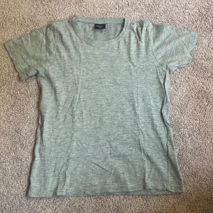 Grå basic t-shirt i bomull - En enkel grå t-shirt från Basic i storlek 158/164. T-shirten har rund hals och korta ärmar, perfekt för chill dagar. Materialet är mjuk bomull och färgen är ljusgrå med en melerad look. Passformen är normal och modellen är klassisk utan extra detaljer.
